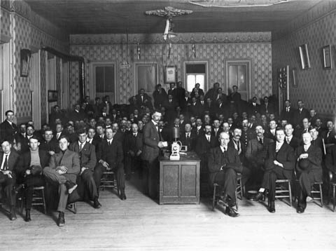 A group of men gather in the Woodmen of the World Hall to view a lantern slide presentation. The contraption on the table is the lantern slide projector.  (WSA Meyers Neg 1009, 1910-1915, photo by Joe Shimitz)